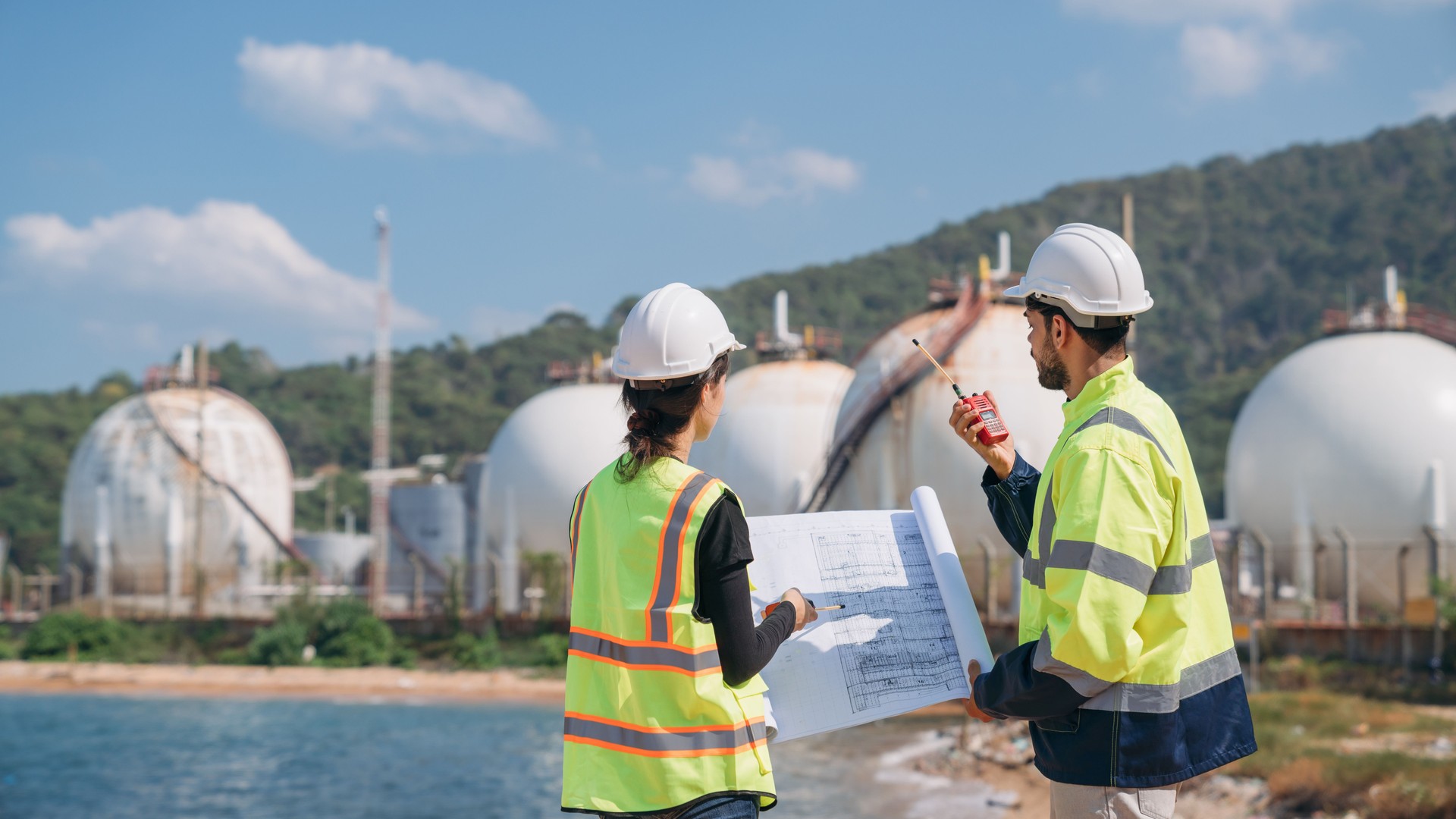 Two engineers in safety gear with blueprints discussing work in front of industrial storage tanks on a clear day.