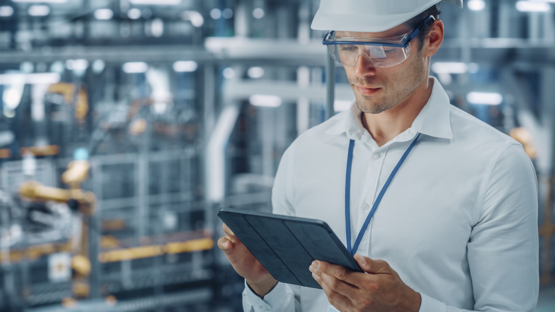 Male Engineer in Safety Goggles and Hard Hat Using Tablet Computer and Looking Out of the Office at a Car Assembly Plant. Industrial Specialist Working on Vehicle Production in Technological Factory.